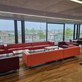 A photo of the RCR. There are 4 red sofas in the middle of a large room arranged facing each other around 3 white coffee tables. The far wall is all floor to ceiling windows . The floor is wooden.
