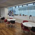 A photograph of the tables in the RCR. here is 3 round, white tables along a wall with 4 red chairs at each. The floor is wooden and the wall behind the tables is white with glass panel in the top half. 